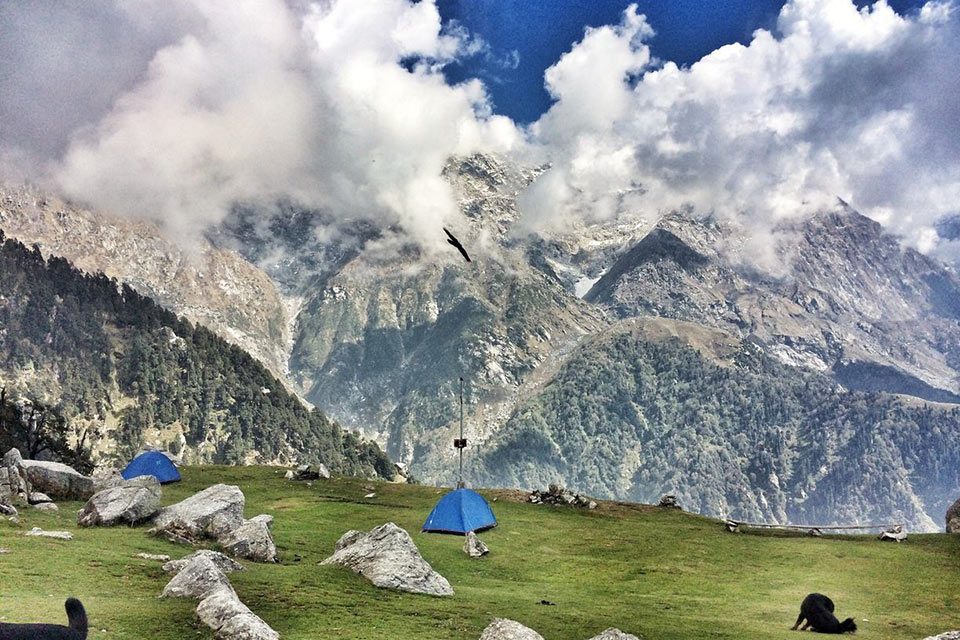 a camping site as seen on a hill at triund with rocky mountains covered in snow in the background